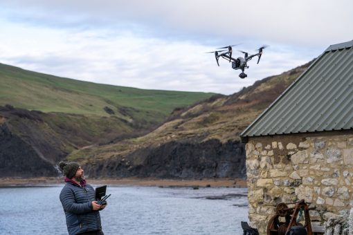 An airborne drone overlooking the side of a cliff and sea. A man is watching the drone and operating it using a drone radio transmitter.