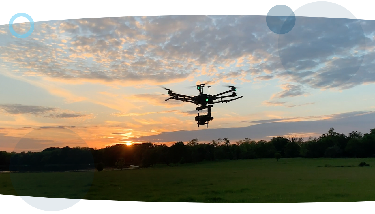 An airborne drone in the middle of a park with trees in the background and the sunsetting behind it. The drone is performing an oil and gas inspection.