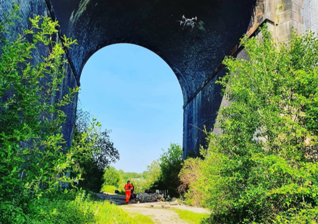 Two RUAS trained pilots walking under a large bridge with an airborne drone performing visual inspections.