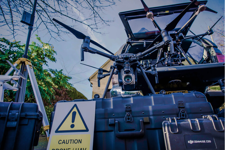 A black drone sat on a drone case with a warning sign leant on it. Setting up drones can be time consuming and dangerous therefore eating into the operators charges.