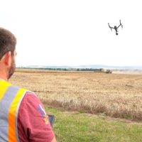 A CAA approved, RUAS trained pilot wearing a high vis vest, flying a drone, using a drone radio transmitter, over a hay field.