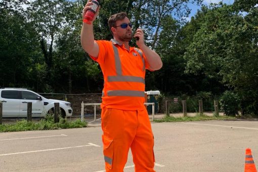 A man stood in a car park, wearing a high vis suit with a walkie talkie in one hand, and an air horn in the other. This signals the take off of the drone during EVLOS training.