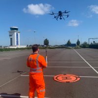 A man wearing a high vis suit with the RUAS logo on it, at an airport. He's flying a Mavic 2 drone, to show the importance of FRZ.
