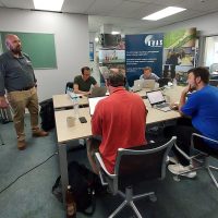 There are six people sat around a table in a RUAS teaching room with laptops, being taught by a RUAS instructor.